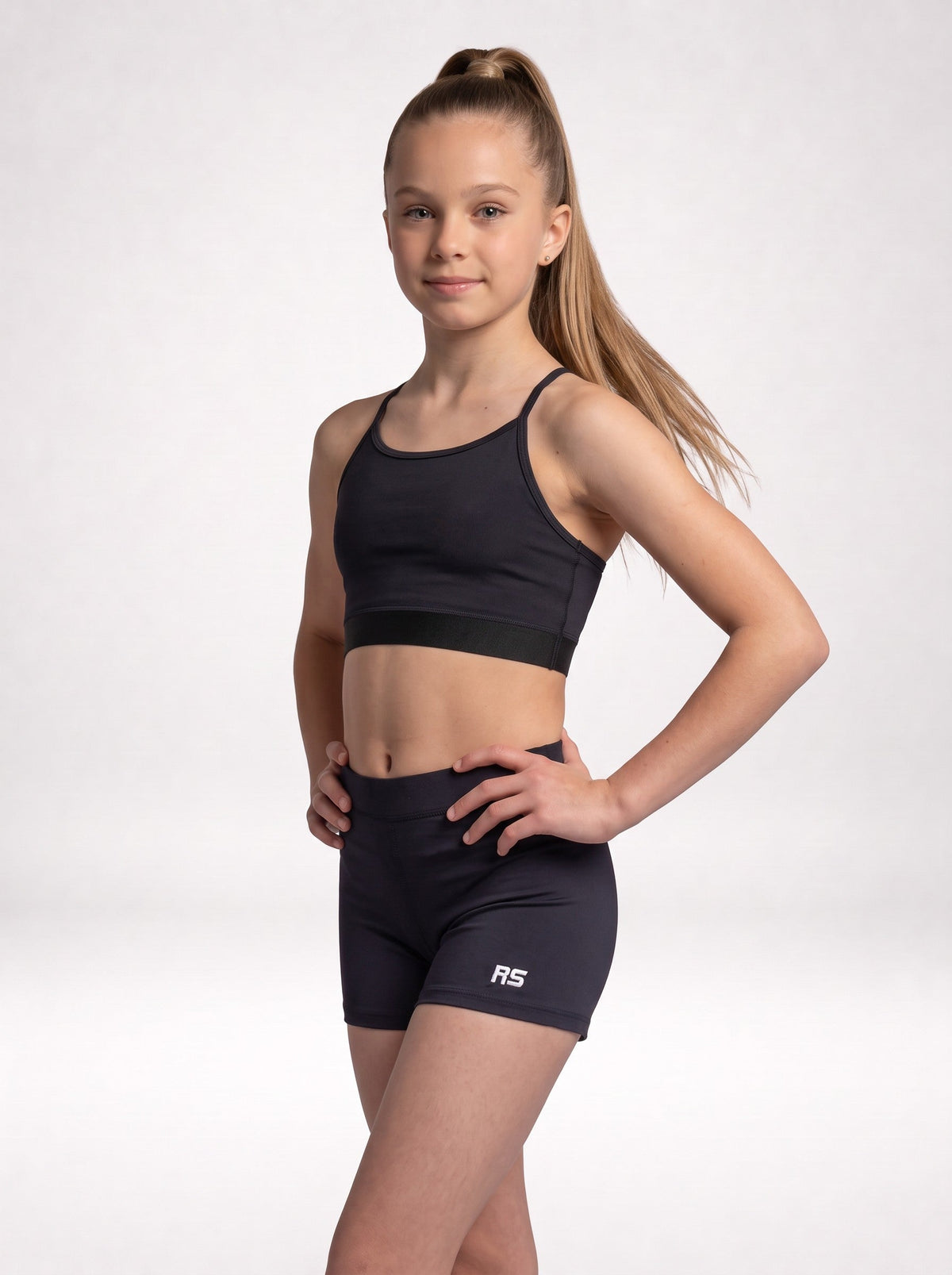 A young girl with a high ponytail stands in a studio wearing a black athletic sports bra and matching black shorts with a white 'RS' logo on the left leg, posed against a light gray background.