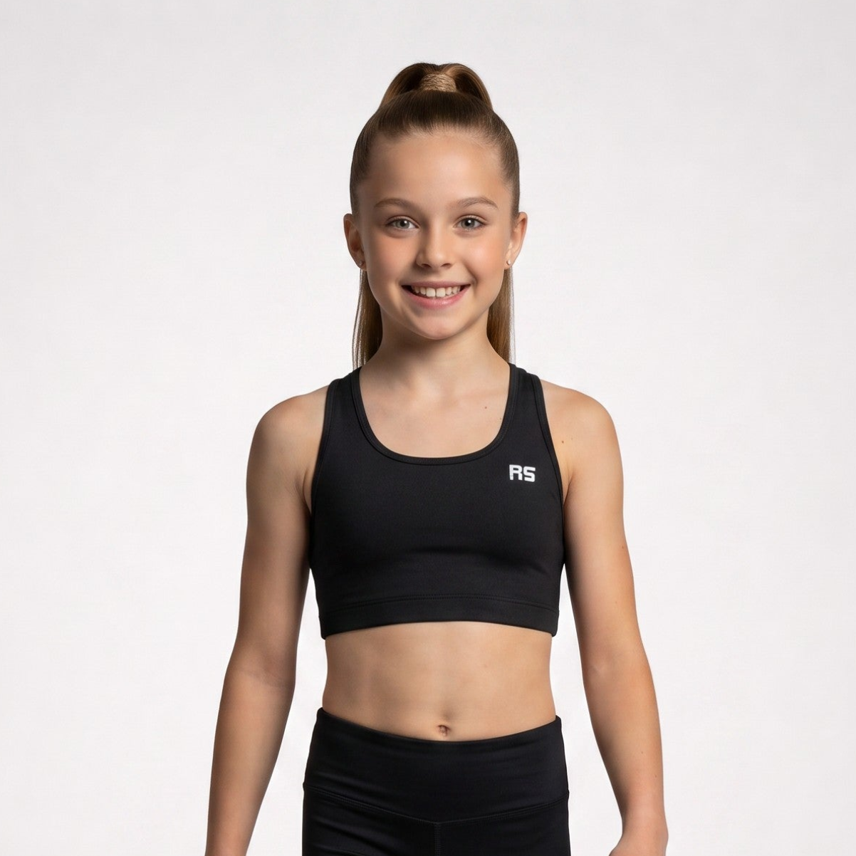 A young girl with her hair in a high ponytail smiles while wearing a black athletic crop top and matching black leggings against a light gray studio background. The crop top features a small white 'RS' logo on the left chest.