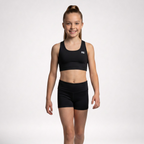 A young girl with her hair in a high ponytail stands against a light gray studio background wearing a black athletic crop top and matching black shorts. A small white logo is visible on the left chest of the top.