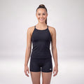 A woman modeling a black athletic tank top and matching black shorts against a light gray studio background. The shorts have a small white logo on the lower left leg.