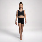 A young girl modeling a black athletic crop top and matching black shorts against a light gray studio background.