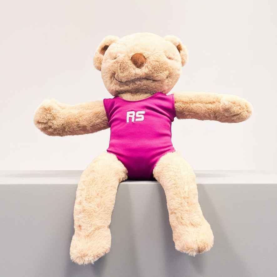 A plush teddy bear sitting on a brown bench, wearing a bright pink sleeveless top with the letters 'RS' in white on the chest.