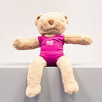 A plush teddy bear sitting on a brown bench, wearing a bright pink sleeveless top with the letters 'RS' in white on the chest.