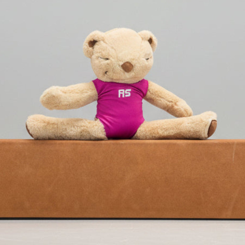 A beige teddy bear sits cross-legged on a brown foam exercise bar, wearing a bright pink one-piece swimsuit with the white letters 'RS' on the chest, set against a white wall and blue carpet.