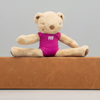A beige teddy bear sits cross-legged on a brown foam exercise bar, wearing a bright pink one-piece swimsuit with the white letters 'RS' on the chest, set against a white wall and blue carpet.