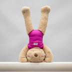 A beige plush teddy bear wearing a bright pink sleeveless bodysuit with the white letters 'RS' on the chest, standing against a plain white wall.