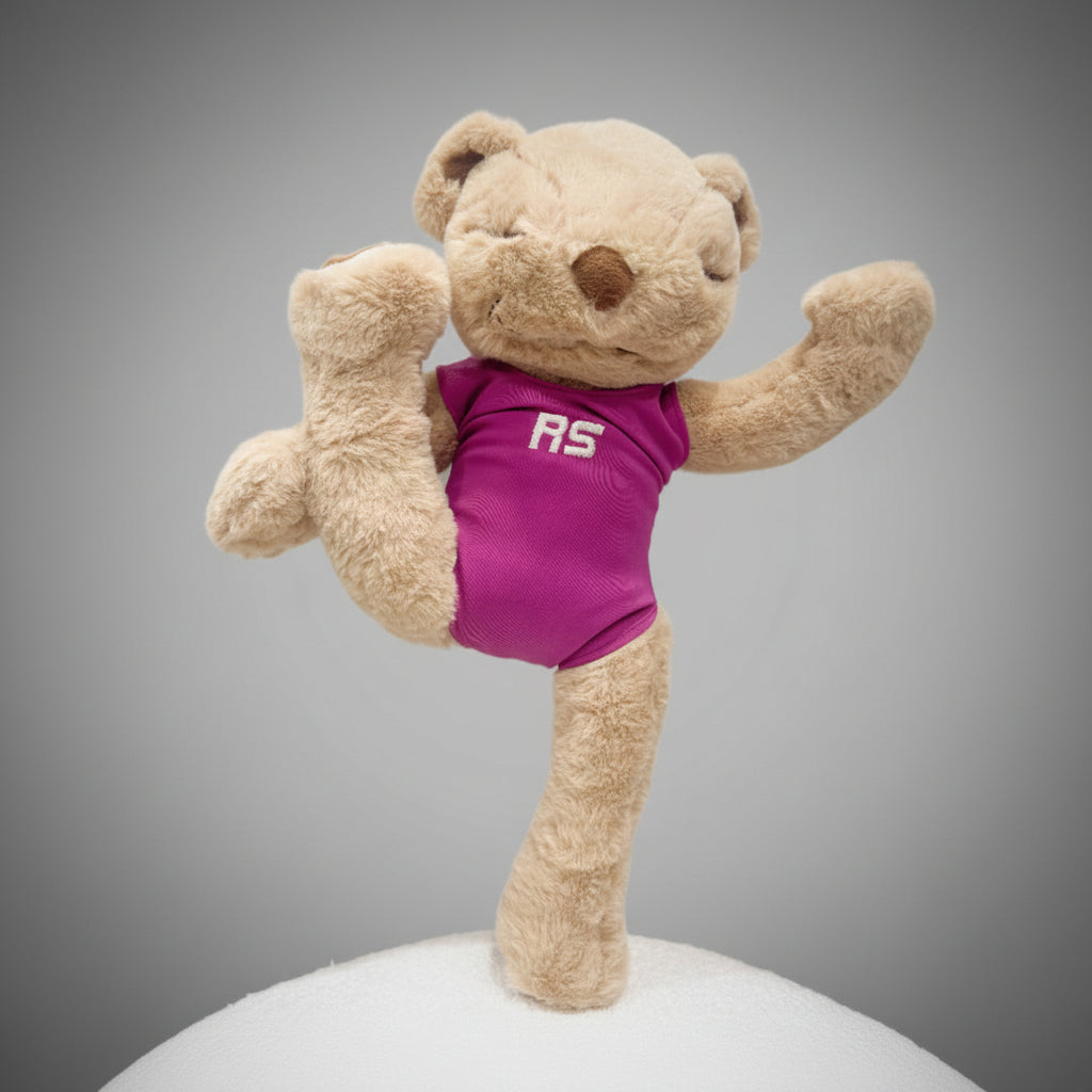 A plush teddy bear toy posed on a white surface against a light gray wall, wearing a bright pink athletic-style top with white 'RS' lettering on the chest.