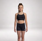 A young girl stands against a light gray studio background wearing a matching black athletic set consisting of a sports bra and fitted shorts. A small white RS logo is visible on the lower right leg of the shorts.