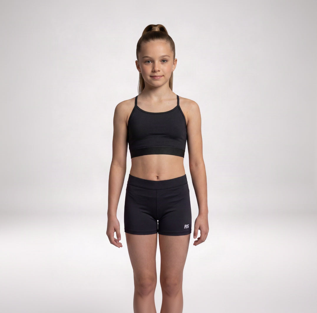 A young girl stands against a light gray studio background wearing a matching black athletic set consisting of a sports bra and fitted shorts. A small white RS logo is visible on the lower right leg of the shorts.