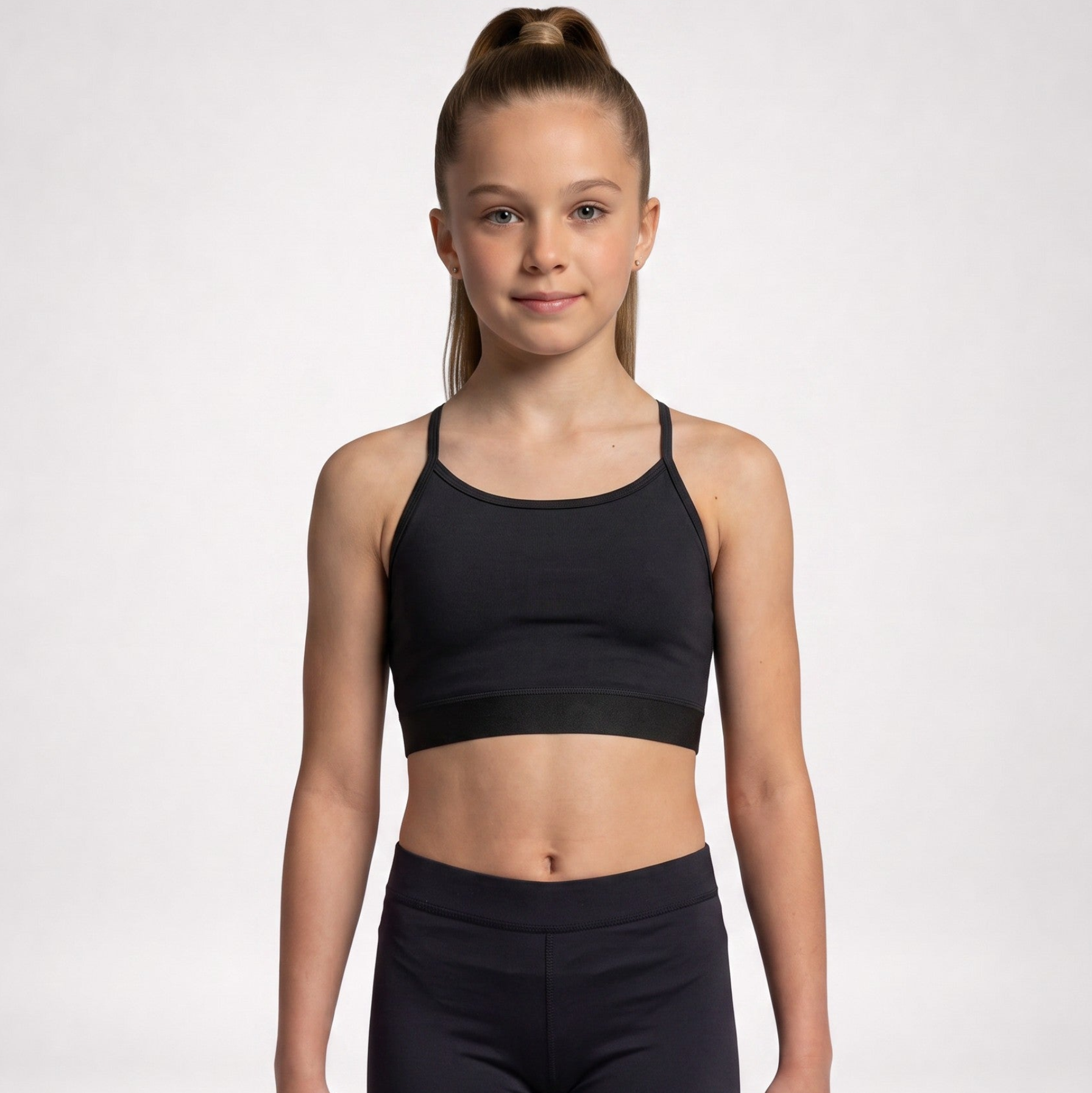 A young girl with her hair in a ponytail stands against a light gray studio background, wearing a black athletic sports bra and matching black leggings.